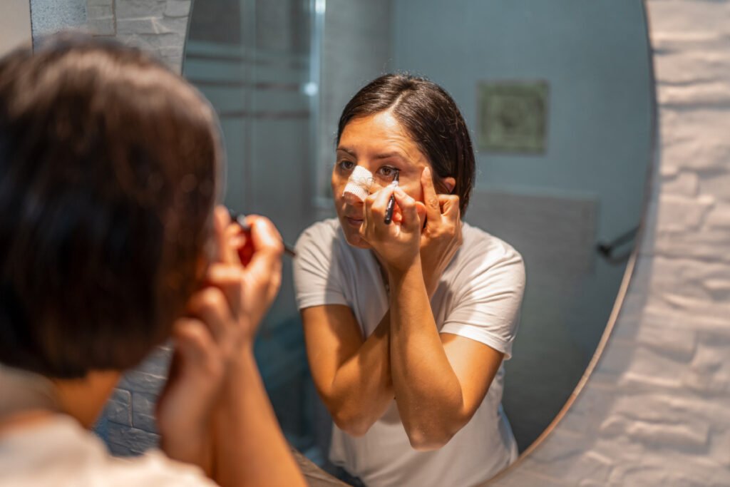 Woman carefully applying makeup in mirror wearing a nose plaster and splint after rhinoplasty surgery