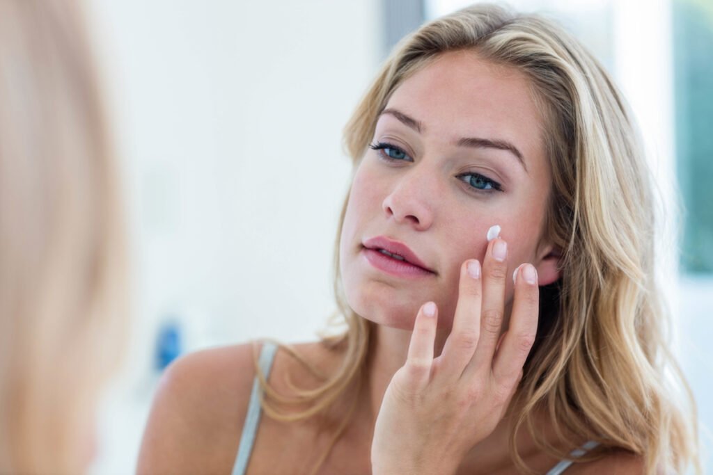 Blonde woman applying sunscreen to face in bathroom mirror